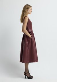 Vestido de imitación de cuero burdeos con un corpiño ajustado, falda acampanada, bolsillos laterales y tirantes. Modelo combinado con tacones negros.