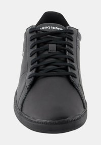 Baskets de sport noires avec une tige en cuir lisse, des lacets plats et une semelle en caoutchouc texturée. Présentent des coutures subtiles et un logo sur la languette.