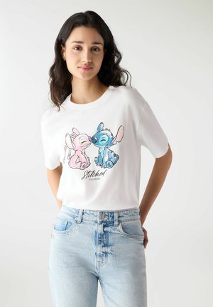 Młoda kobieta w białej koszulce z różowymi i niebieskimi postaciami z kreskówek, siedząca i dotykająca się nosami, w połączeniu z jasnoniebieskimi dżinsami.