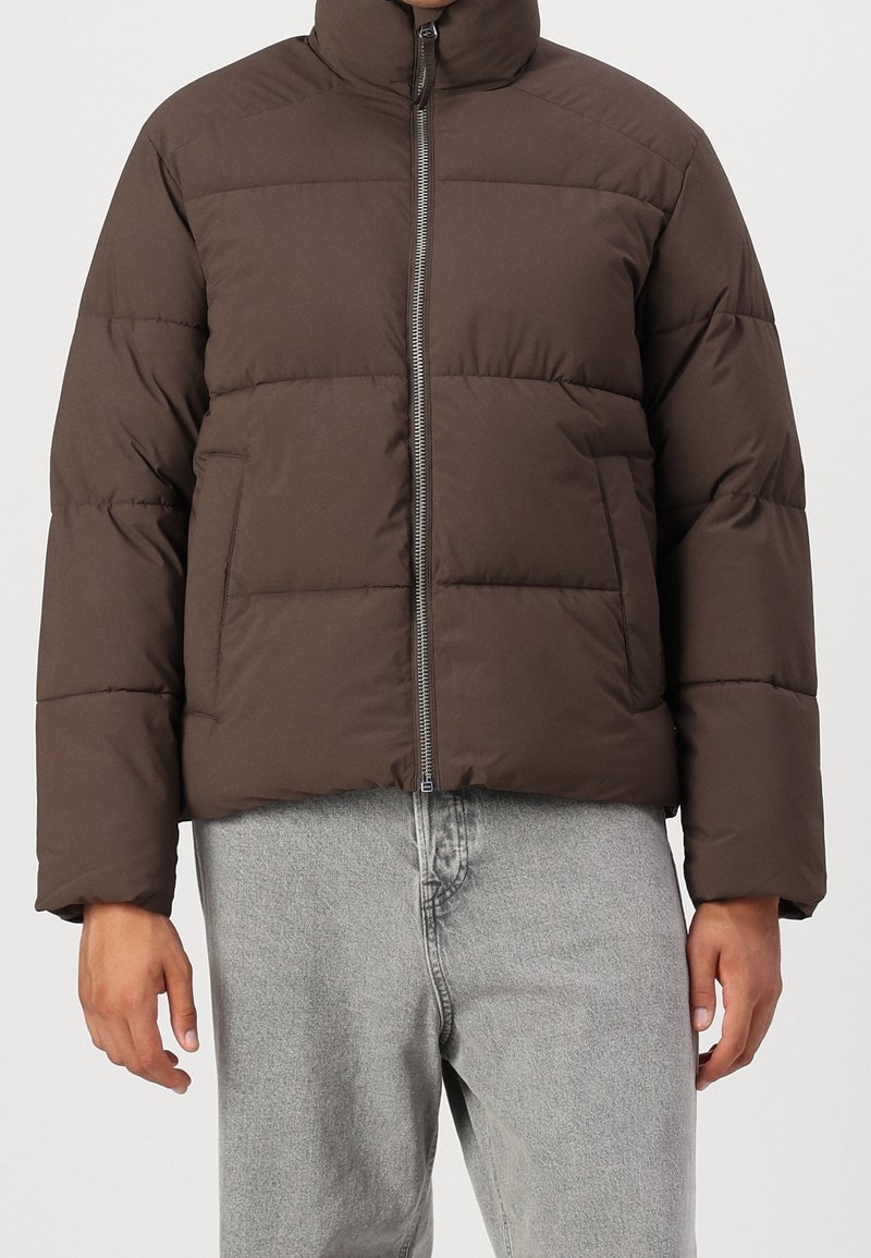 Hnědá prošívaná bunda s vysokým límcem, předním zipem a hranatým střihem. Má prošívané části a je vyrobena z měkké, odolné látky.