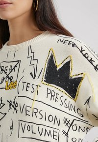 Krämfärgad tröja med svarta tryckta detaljer och livfulla gula accenter. Inkluderar en krondesign och 'Lee'-logotyp, med texturerad sömnad.