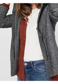Grijs gestructureerd jasje over een roestkleurige gebreide laag, gedragen over een witte top. Voorzien van ritsluiting en knoopdetails op de jeans.
