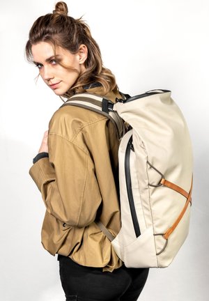 Beige Rucksack mit einer eleganten, weichen Textur, schwarzem Reißverschluss und braunen Lederakzenten. Das Model trägt eine hellbraune Jacke, die das Design der Tasche zur Geltung bringt.