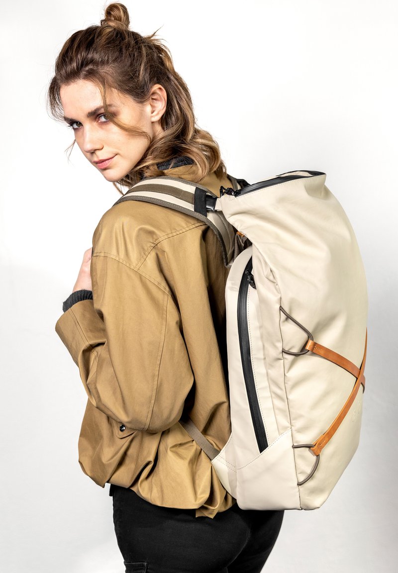Beige Rucksack mit einer eleganten, weichen Textur, schwarzem Reißverschluss und braunen Lederakzenten. Das Model trägt eine hellbraune Jacke, die das Design der Tasche zur Geltung bringt.