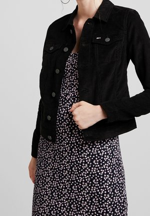 Kobieta ubrana w czarną, zapinaną na guziki kurtkę z sztruksu, na ciemnej sukience w kwiaty, trzymająca ręką dół kurtki, na jednolitym tle.