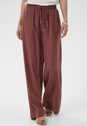 Femme portant un pantalon ample taille haute couleur rouille avec une taille à cordon et des sandales beige à bout ouvert, debout devant un fond blanc.