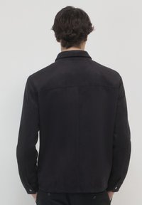 Uomo con i capelli scuri che indossa una giacca nera, in piedi e rivolto verso uno sfondo grigio chiaro uniforme con le mani in tasca.