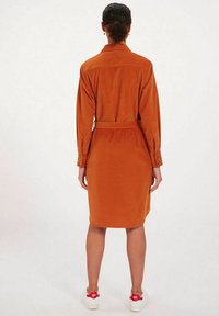 Robe chemise orange brûlé à manches longues, avec des détails de boutons, une ceinture nouée à la taille et une longueur en-dessous du genou, portée avec des baskets blanches.