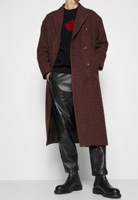 Dubbelknäppt brunt houndstooth-kapp med stora grå knappar, bärs över en svart tröja och svarta läderbyxor, i kombination med svarta ankelskor.