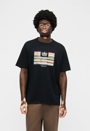 Νεαρός άνδρας με λεύκη στέκεται προς τα εμπρός φορώντας μαύρο μπλουζάκι με σχέδιο και καφέ παντελόνια μπροστά σε απλό ανοιχτό φόντο.