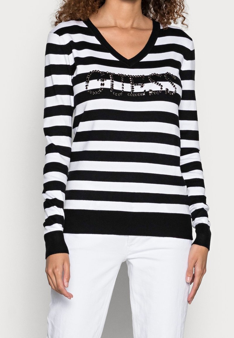 Femme portant un pull à rayures horizontales noires et blanches avec "GUESS" en sequins noirs sur la poitrine et un pantalon blanc.