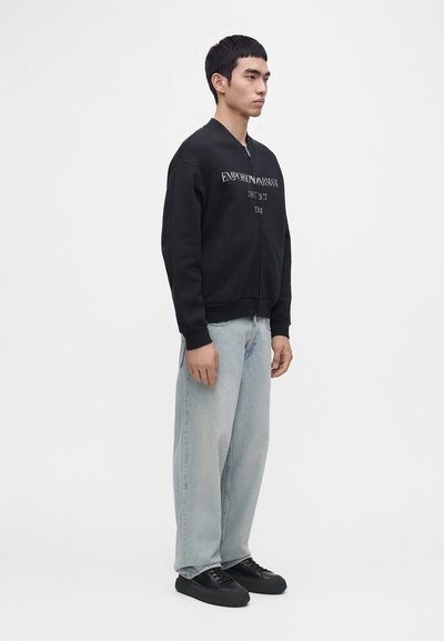 Homme debout portant un sweat à capuche noir zippé avec un texte, un jean large bleu clair et des baskets noires sur fond blanc.
