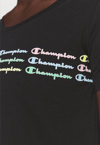 Camiseta negra con múltiples logotipos de "Champion" en colores pastel dispuestos en tres filas horizontales a través del pecho.