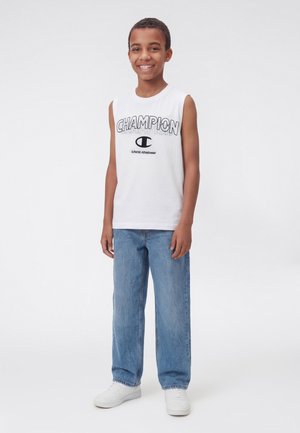 Ragazzo sorridente in piedi, che indossa una maglietta bianca senza maniche Champion, jeans blu e sneakers bianche su uno sfondo chiaro e uniforme.