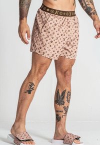 Shorts da bagno beige da uomo con cintura elastica a motivo logo marrone; realizzati in un tessuto leggero e con una vestibilità rilassata. Indossati con sandali coordinati.