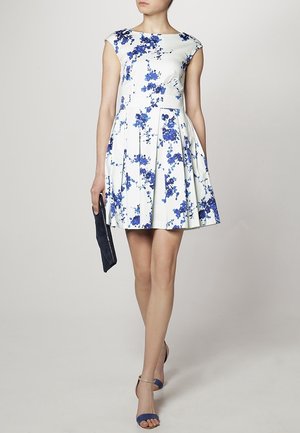 Vestido blanco de mangas de cap sleeve con patrón floral azul, falda acampanada y pliegues. Los accesorios incluyen un bolso de mano azul oscuro y calzado a juego.
