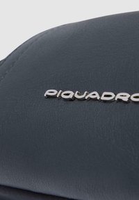 Bolso de cuero gris con una textura suave y un logotipo metálico de Piquadro. El diseño es elegante, con bordes curvados y un hardware mínimo.