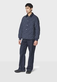 Mørkeblå quilted jakke med kordfløjs krave, trykknaplukning og ribbede manchetter, parret med mørke denimbukser og sorte sko.