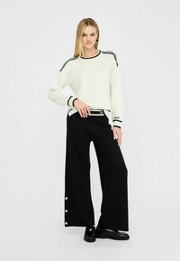 Biały sweter z czarnymi paskami na rękawach i dekolcie, zestawiony z czarnymi, szerokimi spodniami z złotymi guzikami po bokach.