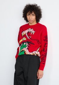 Suéter de punto rojo con un gráfico colorido de un monstruo con colmillos. Mangas largas, cuello acanalado y patrones detallados en negro, rosa y verde.