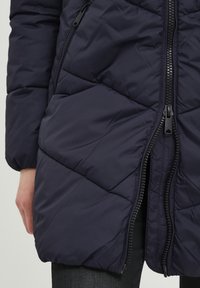 Cămașă umflată de culoare albastru marin cu un model în diamant, fermoar frontal și buzunare laterale. Fabricată dintr-un material neted și ușor.