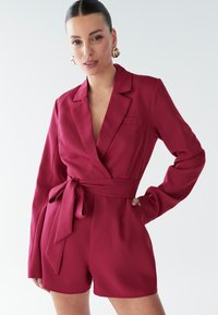Vínový overal s dlhými rukávmi v štýle zavinovacieho kimonového strihu, opasok na zaväzovanie v páse, jedným náprsníkom a hladkou ľahkou textúrou látky.
