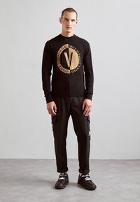 Zwarte trui met een groot beige rond logo met een "V" in het midden, gecombineerd met tapered zwarte cargobroek en zwarte sneakers.