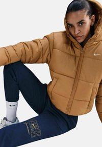 Brązowa kurtka puchowa z kapturem i widocznym przeszyciem. W połączeniu z granatowymi spodniami dresowymi z żółtym logo. Białe skarpetki z czarnym logo.