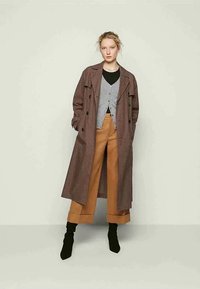 Vrouw staat in een geruite lange jas, grijze cardigan, camel wijde broek en zwarte enkellaarsjes tegen een effen lichte achtergrond.