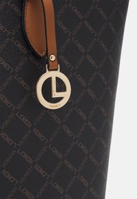 Die schwarze, strukturierte Tragetasche zeichnet sich durch ein sich wiederholendes Logo-Muster in Braun aus. Ein heller Ledergriff mit einem goldenen, runden Logo-Anhänger setzt einen auffälligen Akzent.