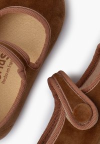 Zapatos Mary Jane de ante marrón con una superficie texturada, que cuentan con una correa de Velcro y un ribete acanalado marrón, con la marca visible en el interior.