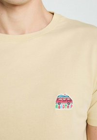 T-shirt en coton beige avec un petit motif brodé représentant une camionnette rouge avec des montagnes, accompagné du texte "ROADTRIP" en lettres colorées.
