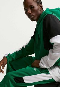 Jeune homme aux cheveux tressés portant un survêtement Nike vert, noir et blanc, assis et regardant devant un fond uni.