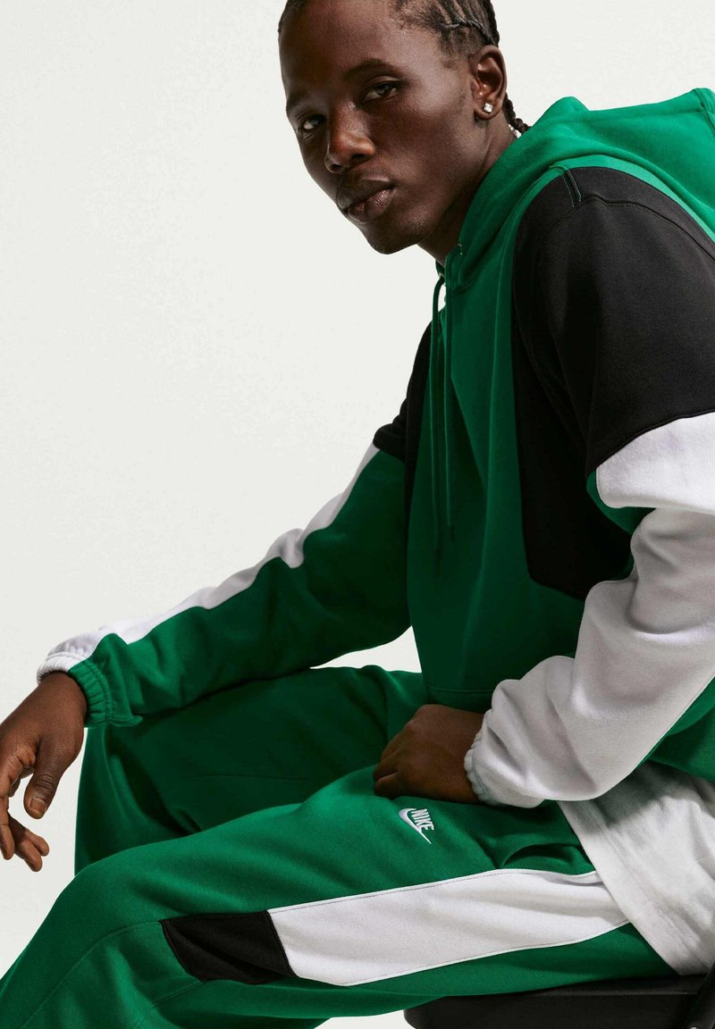 Giovane con capelli intrecciati che indossa una tuta Nike verde, nera e bianca, seduto e che guarda davanti su sfondo uniforme.