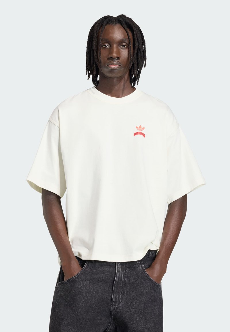 Person mit Dreadlocks, trägt ein übergroßes weißes Adidas-T-Shirt und schwarze Jeans, die Hände in den Taschen, blickt nach vorne.