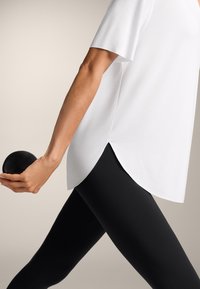 Bílá košile s krátkým rukávem a zaobleným lemem, v kombinaci s černými legínami. Osoba drží černou kouli v pravé ruce.