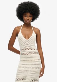 Häkelkleid in Creme mit tiefem V-Ausschnitt, Halterträgern und filigranen offenen Mustern. Die figurbetonte Silhouette betont die Form.