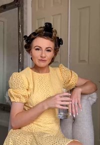 Vestito giallo con motivo a occhio di pernice e maniche a palloncino; bicchiere d'acqua tenuto nella mano destra; capelli in bigodini, seduta su una sedia di tessuto chiaro.
