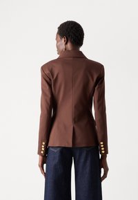 Brun blazer med skreddersydd passform, skarpe rever, gullknappdetaljer og en enkelt splitt bak. Kombinert med mørke denimbukser.