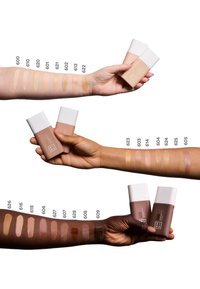 Tre sett med armer viser ulike foundationnyanser i flasker med hvite hetter. Prøvene varierer fra lyse til mørke toner, merket med tall.