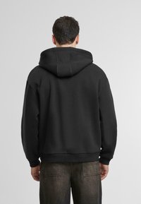 Crna dukserica s kapuljačom, opuštenog kroja, s rebrastim rukavima i donjim rubom. Glatka tekstura i minimalistički dizajn bez vidljivih logotipa.