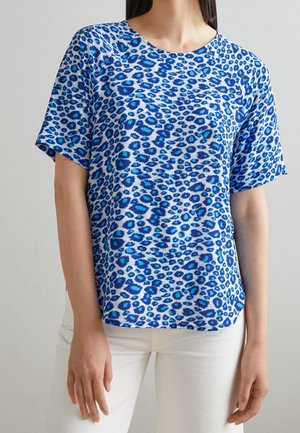 Femme portant une chemise à manches courtes bleue et blanche à motif léopard avec un col rond, associée à un pantalon blanc.