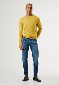 Pull en tricot jaune, jeans slim-fit bleu et bottines noires. Texture lisse, design épuré. Le mannequin se tient avec assurance contre un fond clair.