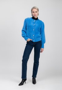 Frau in einem blauen, flauschigen, knöpfbaren Cardigan über einem schwarzen Rollkragenpullover, dunklen blauen Jeans und schwarzen, absatzlosen Stiefeletten, die vor einem einfarbigen Hintergrund steht.
