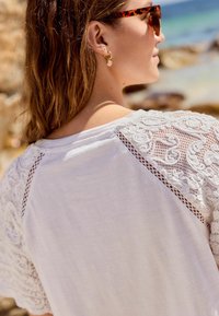 Camicia bianca di cotone con dettagli in pizzo sulle spalle e un sottile motivo floreale. Il modello indossa orecchini rotondi e occhiali da sole.
