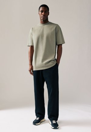 Mann steht mit Händen in den Taschen, trägt ein übergroßes hellgraues T-Shirt, schwarze Hose und schwarze Sneakers vor hellem Hintergrund.