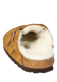 Tan suedne copatice s mehkimi belimi krznimi podlogo, z vrvico iz plute in bakrenim detajlom na strani. Oblikovanje z odprtimi petami za enostavno obuvanje.