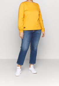 T-shirt giallo a maniche lunghe in cotone, abbinata a jeans di denim blu medio e sneakers bianche. Presenta un piccolo etichetta del brand sulla maglietta.