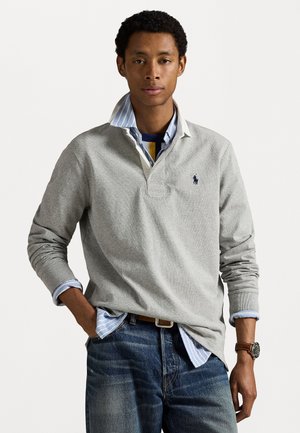 Grå bomullspolo med blå detaljer, lagd over en lyseblå stripete skjorte. Mørkeblå jeans og et brunt klokke er synlige.