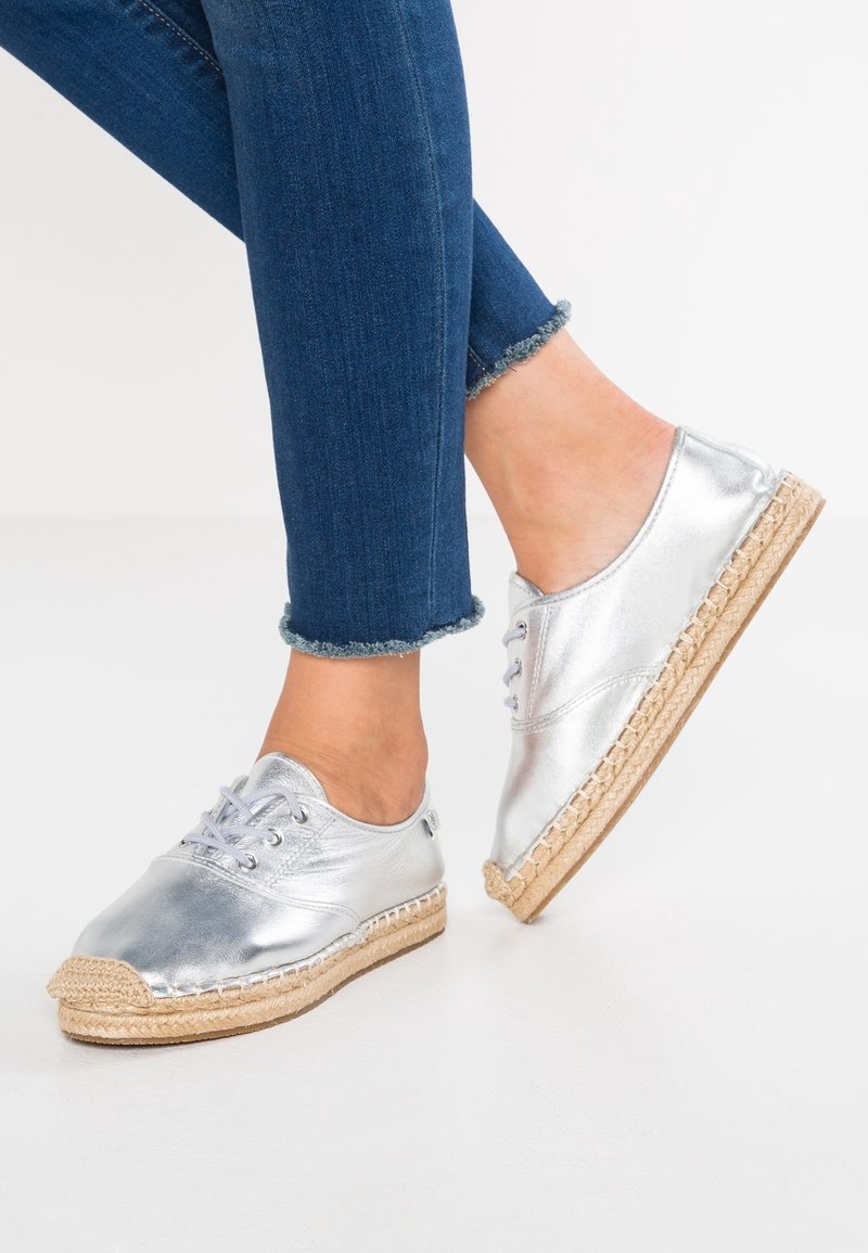 Espadrilles métalliques en argent avec un design à lacets et une semelle en jute, associées à un jean en denim bleu foncé retroussé.